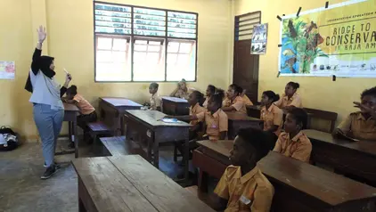 Einblick in ein Klassenzimmer in Indonesien: Eine Lehrerin steht vor der Klasse und hebt den Arm. Die Kinder sitzen auf Holzbänken und folgen der Lehrerin.