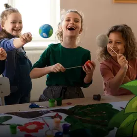 Das Bild zeigt 4 Kinder, die Plakate malen  - im Comic Stil hinzugefügt - eine grüne Pflanze mit einer Schnecke darauf unten rechts.