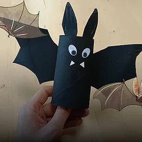 Eine aus Pappe gebastelte Fledermaus wird hochgehalten - im Comicstil hinzugefügt - zwei fliegende Fledermäuse.