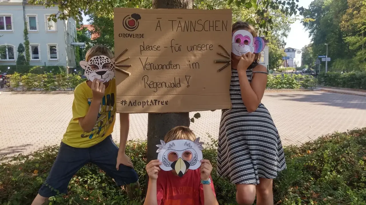 Drei Kinder posieren vor einem Schild an einem Baumstamm. Darauf steht ein Text in einem Dialekt. 