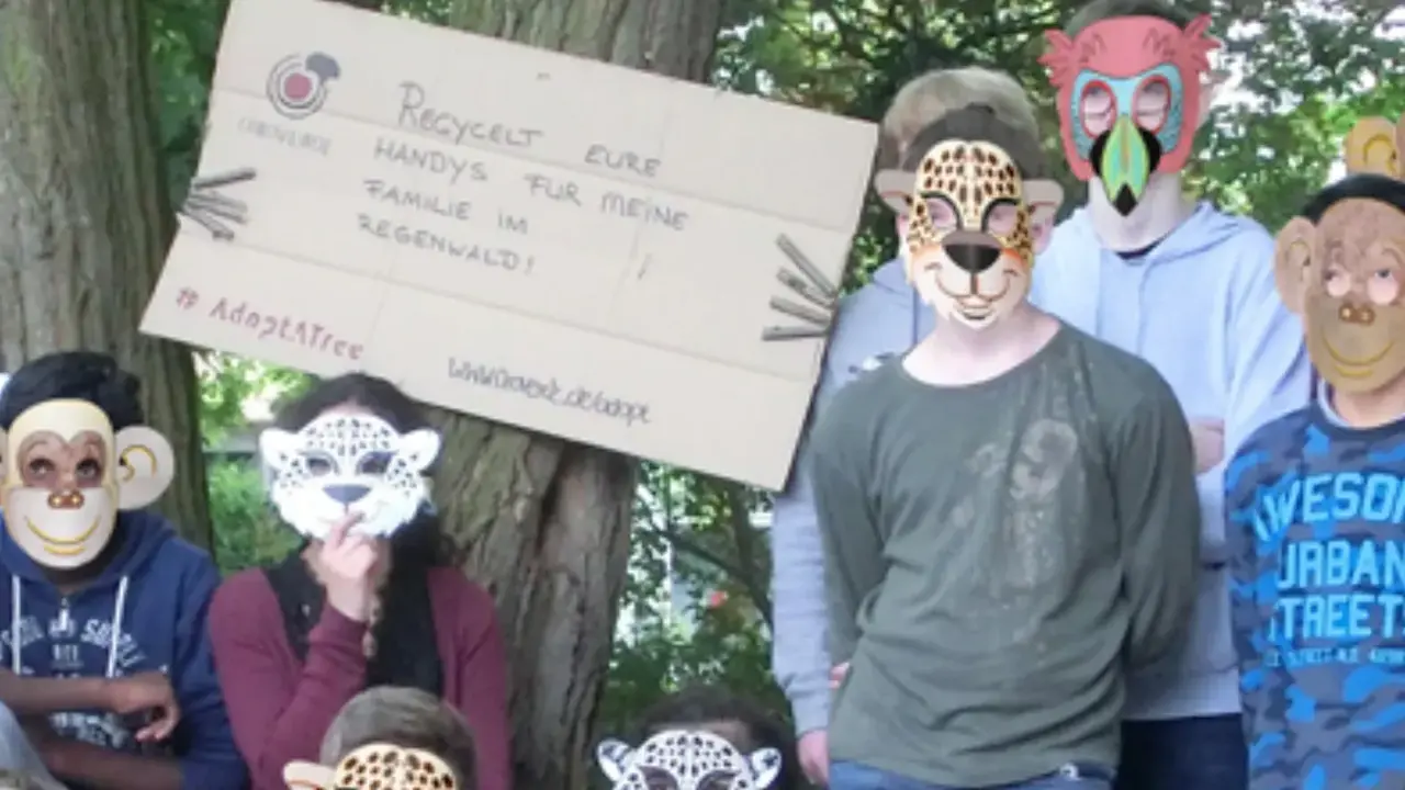 Das Bild zeigt einige Schüler*innen, die vor einem Baumstamm stehen. Der Stamm hält ein Schild mit Slogan und die Kinder Tiermasken. 