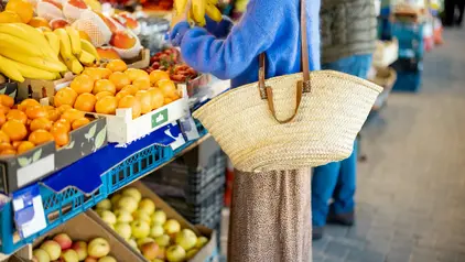 An einem Marktstand, der Obst anbietet, steht eine Frau mit einem Einkaufskorb.