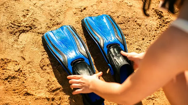 Schwimmflossen ©Kateryna Hliznitsova via Unsplash+ Zu sehen sind 2 Hände, die zwei blau/schwarze Schwimmflossen im Sand abstellen.