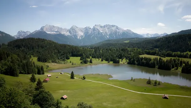 Landschaft mit Wald, Wiese einem See und im Hintergrund Bergen.