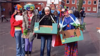 Vier Mädchen stehen auf einem Schulhof und tragen bunte Masken aus Federn. Sie haben einen Bauchladen umgehängt. 