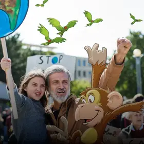 Ein Kind auf dem Arm eines Mannes mit einem Schild in der Hand bei der Demonstration - im Comic Stil hinzugefügt - ein Affe mit erhobenen Händen und fliegende Vögel im Hintergrund.