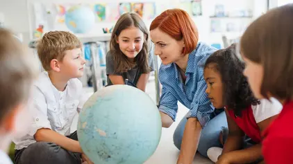 Das Bild zeigt eine Gruppe Kinder mit einer Lehrerin, die sich über einen Globus beugen. 