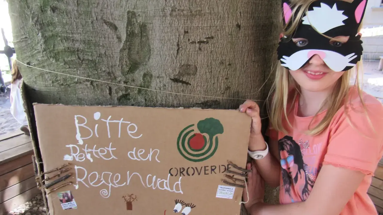 Ein Mädchen steht neben einem Baumstamm und hält ein Schild mit der Aufschrift: "Bitte rettet den Regenwald".