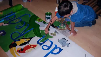 Ein Junge sitzt auf dem Boden und bemalt ein farbenfrohes Schild mit großen blauen Buchstaben. 