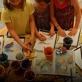 Kinder basteln an einem Tisch mit Papier - im Comic Stil hinzugefügt - mehrere Käfer und ein Äffchen.