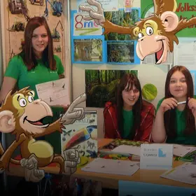 Eine Gruppe Mädchen an einem Infostand - im Comic Stil hinzugefügt - ein Affe, der auf Plakate zeigt. 